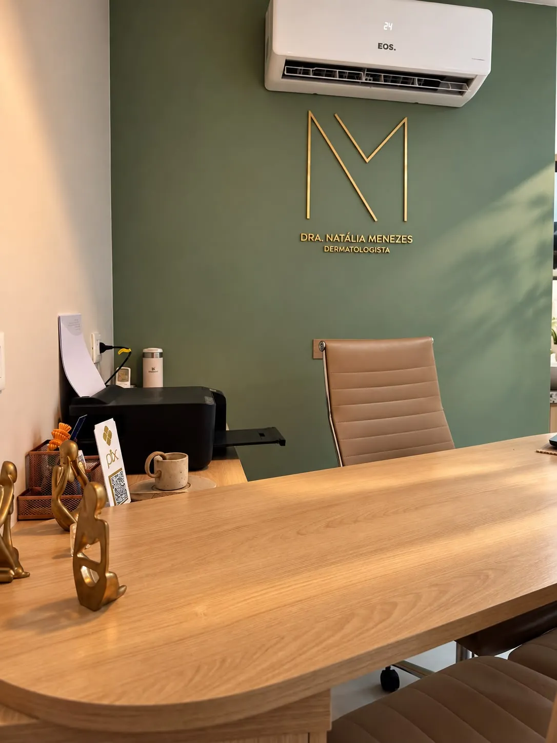 Mesa de atendimento do consultório, com a parede verde e o logo da Dra. Natália Menezes